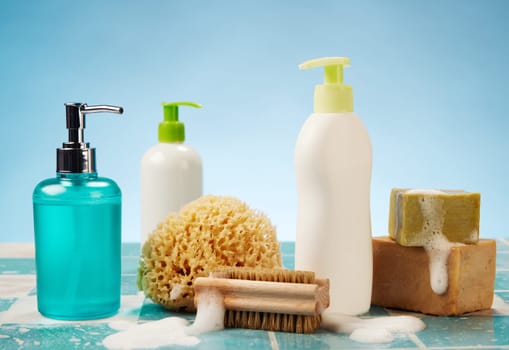 Bottles with dispensers placed near foamy soap and zero waste brush with sponge on wet tiled surface against blue background