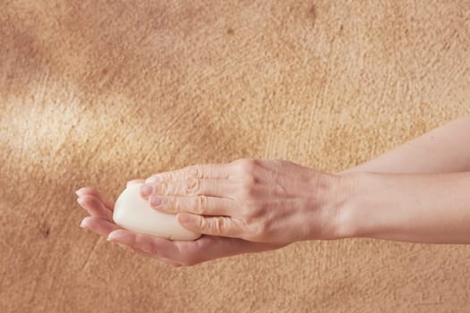 Womans hands holding white piece of soap on mediterranean brown or beige background for spa and wellness concept