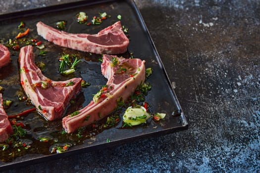 From above of uncooked lamb chops placed on black baking pan with greens and oil on table in light kitchen
