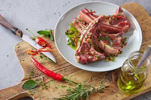 From above of plate with raw lamb chops placed on wooden chopping board with greens and hot pepper in kitchen