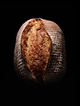Top view of whole brown loaf of bread with crispy crust covered with flour placed on black background in dark studio