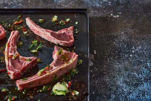 From above of uncooked lamb chops placed on black tray with chopped greens and oil on dark table in light kitchen