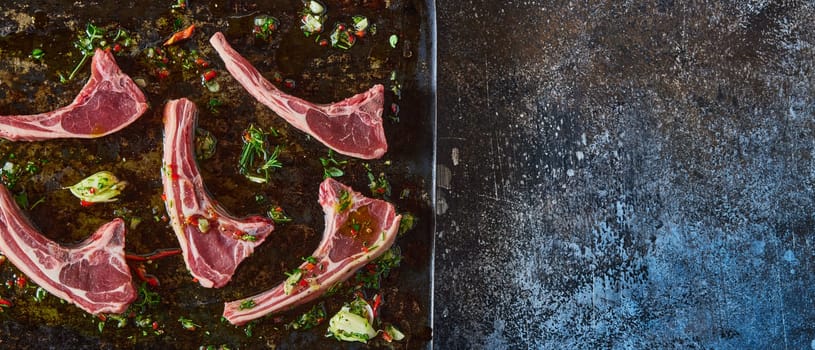 Top view panorama of raw lamb chops placed on metal black baking pan with greens on dark background in kitchen