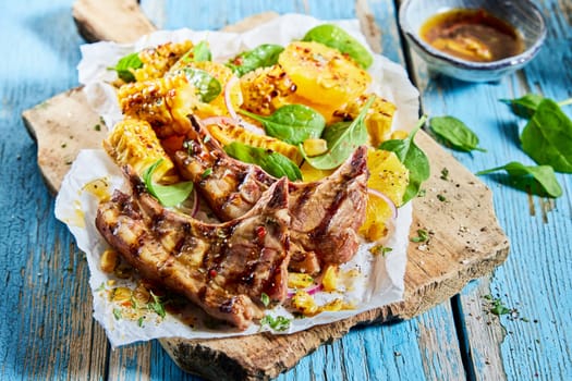 Delicious grilled lamb chops with sliced corn served on wooden cutting board with spinach leaves and orange circles on shabby table in light kitchen