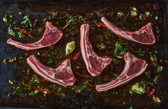 Top view of uncooked fresh lamb chops placed on dark surface with oil and greens in light kitchen during cooking process