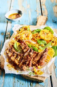 Tasty grilled lamb chops with corn and orange slices served with spinach leaves on cutting board on old blue wooden table in light kitchen