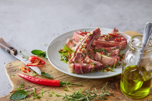 Plate with raw marinated lamb chops on wooden chopping board with oil and chili pepper on gray background in kitchen