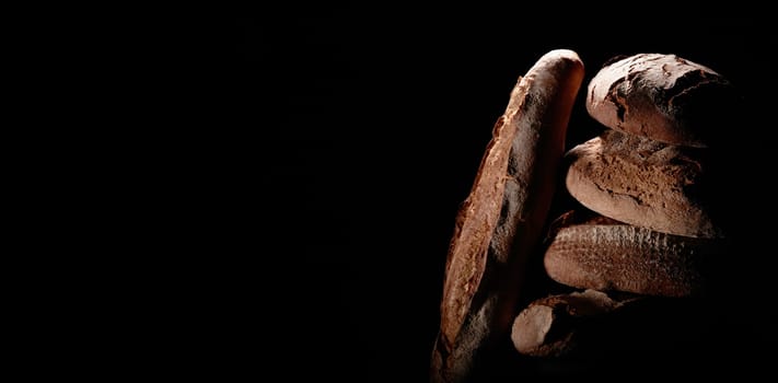 Bunch of delicious freshly baked bread loaves with baguette stacked on top of each other on black background in dark studio