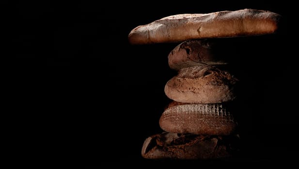 Bunch of delicious freshly baked bread loaves and baguette stacked on top of each other against black background in studio