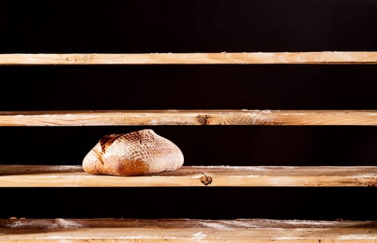 Loaf of baked bread with crust placed on empty wooden shelves in bakery on black background during scarce poverty time in bakery