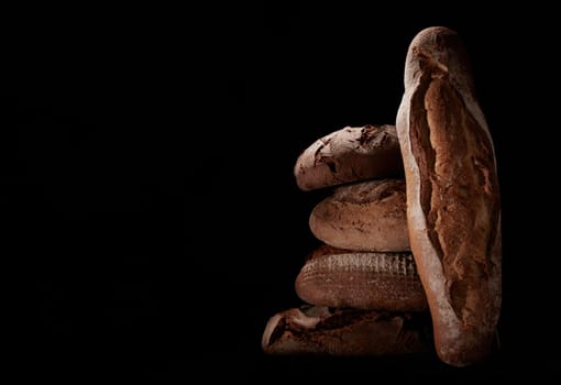Heap of assorted brown freshly baked loaves of bread and baguette stacked on black background in studio with dim light