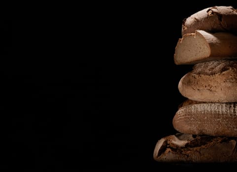 Heap of freshly baked loaves of bread with crust stacked on each other against black background in studio with obscure light