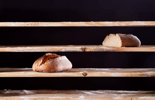 Two baked loaves of bread placed on empty wooden shelves with flour of bakehouse on black background during war time