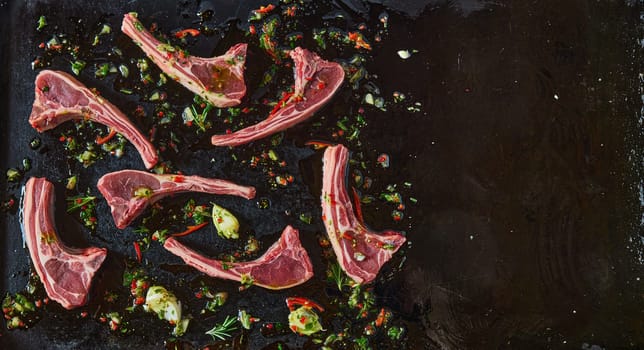 Top view of uncooked lamb chops with chili pepper on black background with chopped greens and garlic cloves in kitchen