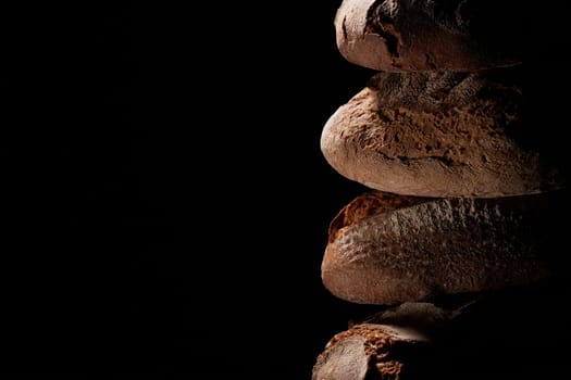 Heap of fresh baked loaves with crispy crust stacked on top of each other against black background in studio with dim light
