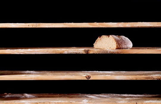 Half of bread loaf placed on empty wooden shelf with white flour on black background during war time in bakery