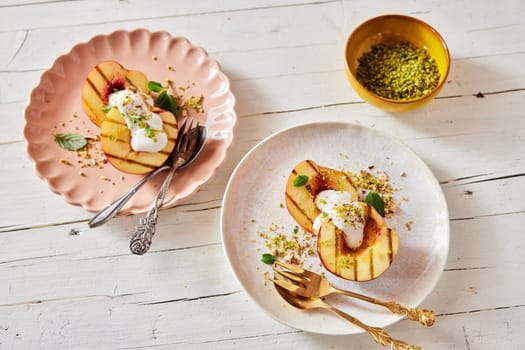 Top view of sweet grilled halves of ripe peaches with yogurt and mint sprinkled with pistachios served on plates on wooden table