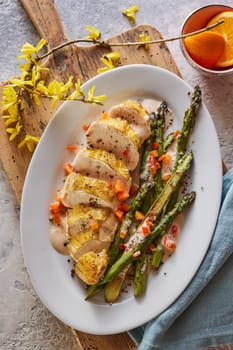 Top view of plate with delicious slices of chicken breast served with asparagus and carrot on wooden cutting board with spring flowers