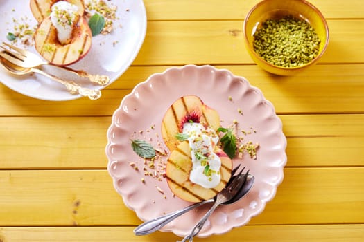 Overhead view of plate with delicious sweet nectarines with yogurt and mint leaves served on wooden table with bowl of pistachios