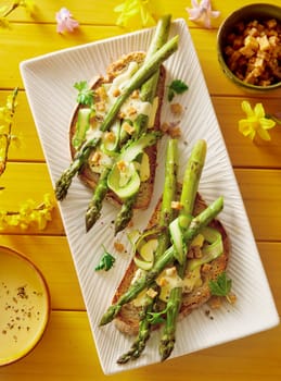 Top view of yummy green toasts with fresh asparagus and cucumber served on plate on table with spring flowers