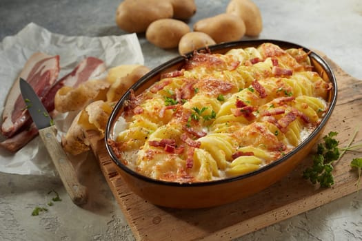 From above of appetizing potato gratin with cheesy sauce prepared in casserole pan and decorated with herbs and bacon slices served on wooden cutting board