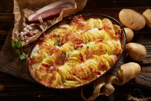 From above of homemade traditional casserole with sliced potato and cheesy cream served in oval pan with bacon pieces and parsley on wooden cutting board
