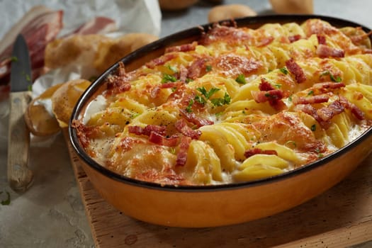 From above of yummy casserole with sliced potato bacon and cheese in oval pan placed on wooden cutting board near knife and peel in kitchen