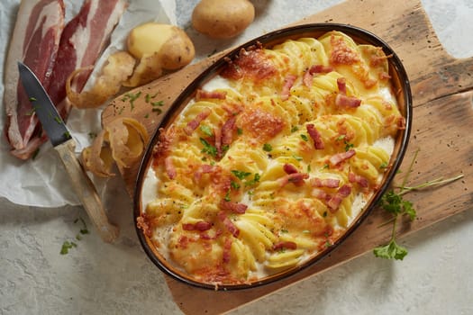Top view of oval pan with appetizing potato gratin with cheese and bacon served on wooden cutting board on table in kitchen