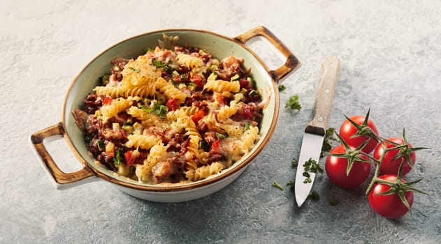 High angle of ceramic baking dish with fusilli pasta placed near bunch of tomatoes and knife with chopped herbs