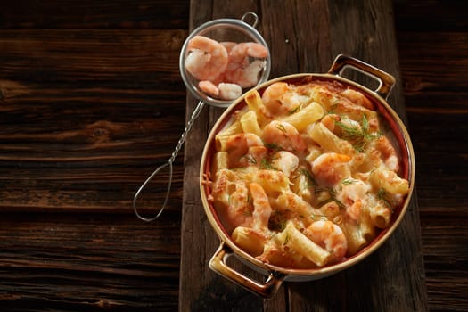 Top view of saucepan with appetizing creamy rigatoni pasta with shrimps and herbs served on wooden cutting board in kitchen
