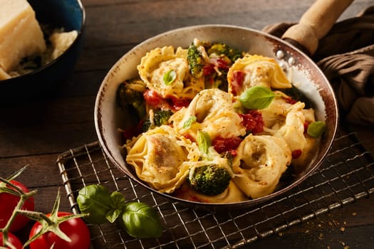 High angle of appetizing homemade tortelloni pasta with broccoli and tomatoes on metal grill near cheese in bowl on wooden table