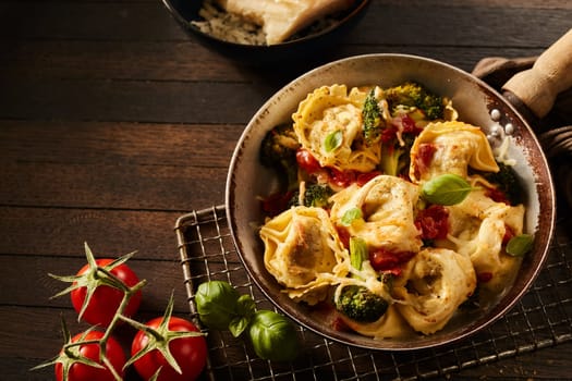 High angle of yummy Italian tortelloni with broccoli and tomatoes in hot frying pan on metal grid near parmesan cheese on wooden table
