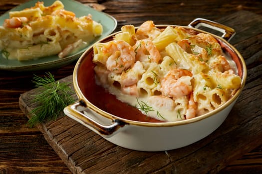 High angle of delicious rigatoni pasta and shrimps gratin baked in ceramic casserole pan with cream sauce and served in plate on wooden table
