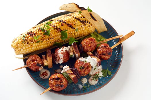High angle of tasty grilled corn cob and meatballs on sticks with sauce on ceramic plate on white background