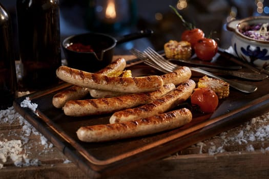 High angle of appetizing homemade grilled corn sausages and tomatoes on wooden tray with sauce cabbage salad near bottles of beer at winter night
