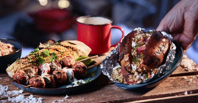 Crop anonymous person taking bowl with sweet potato with vegetables and bulgur from table with BBQ meatballs and corn at winter night