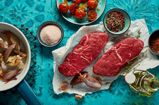 Top view of uncooked striploin beef steak on baking parchment served with cups of pepper condiment near bottle of oil and onion