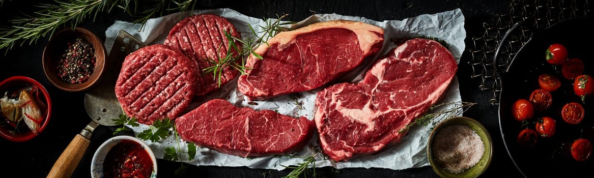 Overhead of raw beef sirloin and ribeye steaks and patties arranged with spices and pan with grilled tomatoes