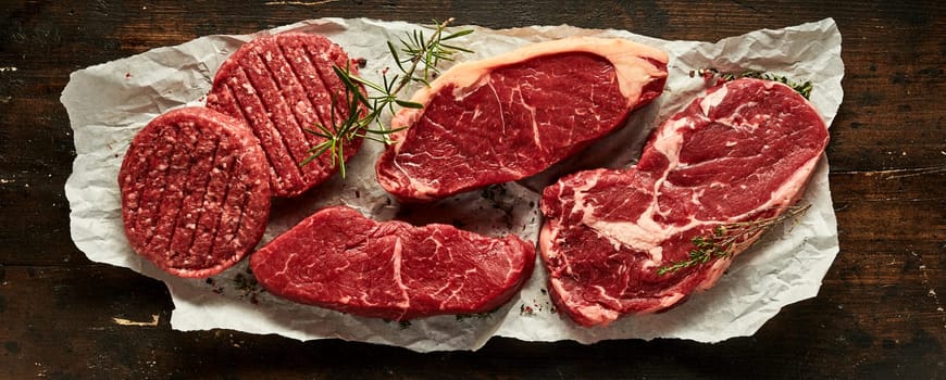 Top view of raw beef sirloins and ribeye steaks with burger cutlets and rosemary twig on creased paper on wooden table