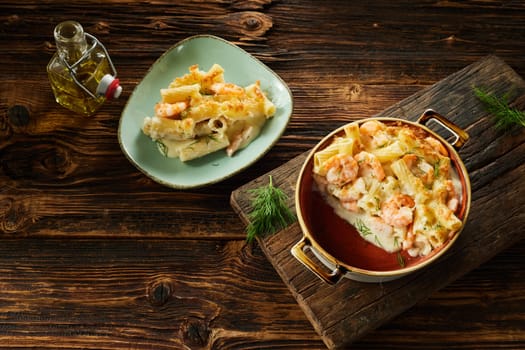 Top view of tasty homemade gratin with rigatoni pasta and shrimps served on wooden table in ceramic casserole pan and plate with bottle of olive oil