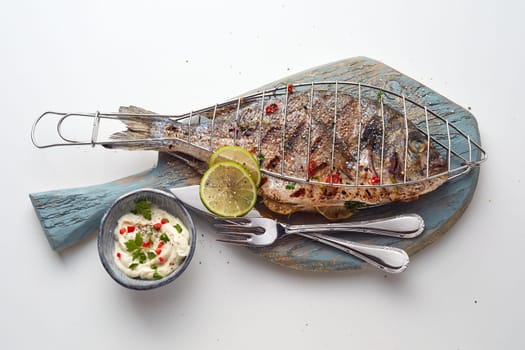 High angle of tasty grilled fish with limes served on wooden cupboards placed on light table near bowl with sauce