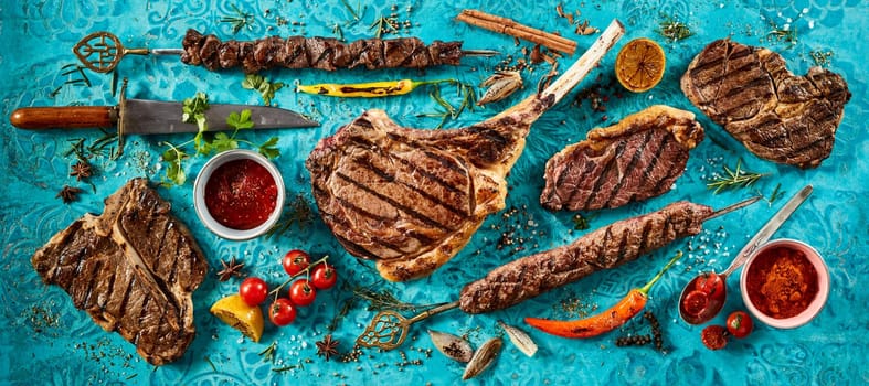 High angle of tasty grilled meat steaks and spicy sauces placed on blue table with fresh tomatoes