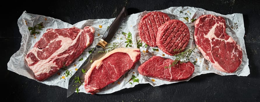Top view of various uncooked ribeye and sirloin beef steaks arranged with burger cutlets and sharp knife seasoned with herbs