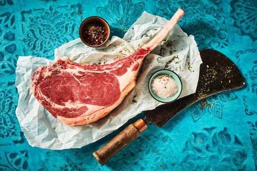 Top view of ribeye served on long rib with cup of salt and pepper on paper and chopping ax on blue table