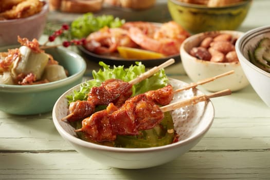 Tasty grilled chicken skewers in bowl with lettuce leaf against various appetizers on table on blurred background
