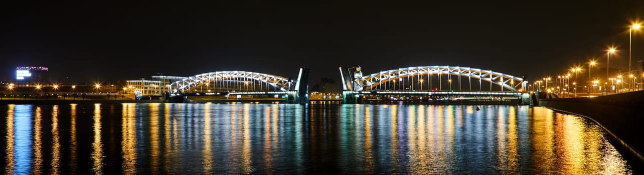 Panoramic view of illuminated arched drawbridge with luminous lights reflecting on river at night in Saint Petersburg