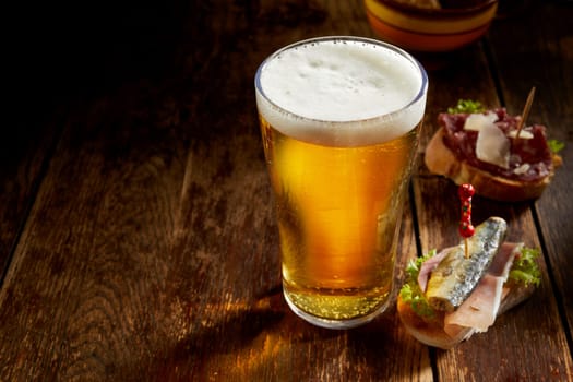 Pint of cold draft beer in a tall glass with delicious assorted tapas on fresh baguette served on a rustic counter with copyspace in a pub or tavern