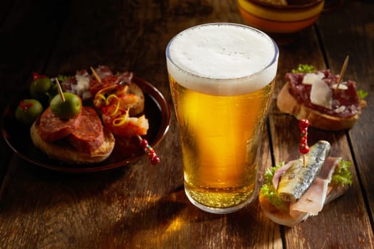 Tasty selection of savory Spanish tapas served with a cold pint of beer with frothy head on a rustic shadowy wooden counter in a tavern in a high angle close up view