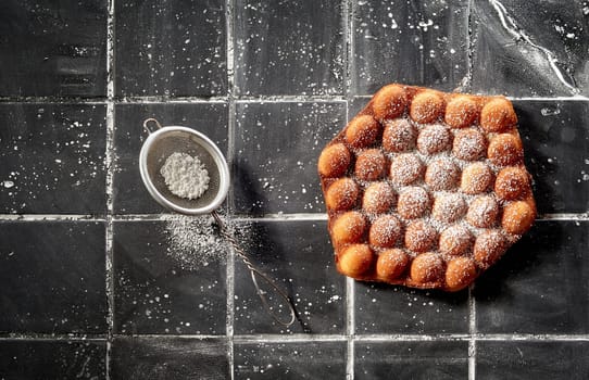 Trendy crispy golden bubble or egg waffle sprinkled with icing sugar in a top down view on dark grey tiles with copyspace