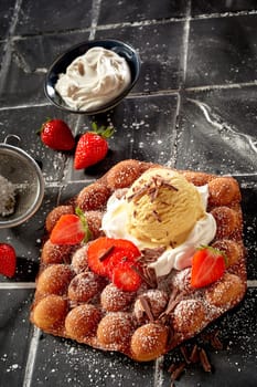Delicious bubble waffle with ripe strawberries and whipped cream topped with vanilla ice cream drizzled with chocolate flakes and icing sugar for a gourmet summer dessert